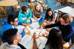 Diverse group of teen high school students are sitting around round table in library. They are studying books and papers to prepare for a test. Caucasian, African American, Hispanic, and Indian teen boys and girls are smiling while working together on class project.